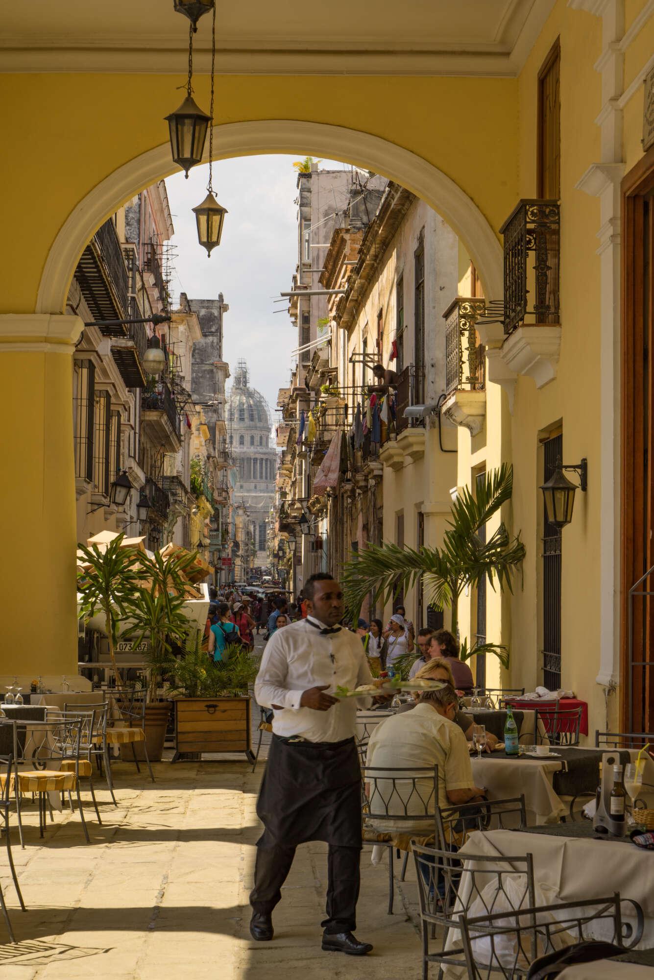 We are under the balcony of one of the old houses in Havanna, turned restaurant. We see a waiter switfly walking inside to, no doubt, pick up some orders for the customers. Beyond the restaurant's terrace, we look down a colourfull Havanna street, where, in the distance, our eye meets the capitol building of Havanna. We are under the balcony of one of the old houses in Havanna, turned restaurant. We see a waiter switfly walking inside to, no doubt, pick up some orders for the customers. Beyond the restaurant's terrace, we look down a colourfull Havanna street, where, in the distance, our eye meets the capitol building of Havanna.